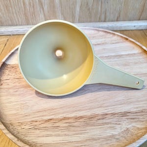 May include: A vintage, pale yellow Tupperware funnel with a handle and a small hole at the bottom. The funnel is resting on a round, wooden serving tray. The word "Tupperware" is printed on the handle.