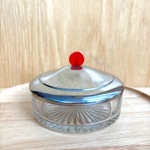 May include: A clear glass jar with a silver lid and a red knob on top. The jar has a decorative, fan-like pattern on the bottom half. The lid has a silver rim. The jar is sitting on a wooden surface.