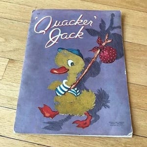 May include: A vintage children's book cover featuring a cartoon duck wearing a blue and white striped shirt and a red cap. The duck is carrying a stick with a red polka dot bag attached to it. The book title is "Quack'er Jack" in a whimsical font.