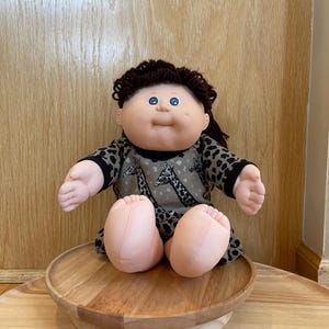 May include: A vintage doll with brown curly hair and blue eyes, wearing a gray and black patterned dress with giraffe illustrations. The doll is seated on a wooden pedestal, with its arms and legs extended.