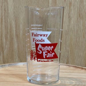 May include: A clear glass measuring cup with red and white text that reads "Fairway Foods" and "Super Fair". The cup has measurement markings and a spout for easy pouring. The design includes a Ferris wheel and other fairground imagery.