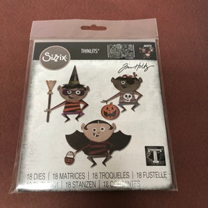 May include: A Sizzix Thinlits die-cut set with a Halloween theme. The package shows three cartoon characters: a witch, a pirate, and a vampire. The witch has a broom, the pirate an eye patch, and the vampire a basket. The set contains 18 dies for crafting.