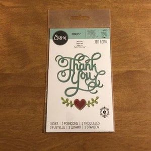 Peut inclure: Un paquet de matrices de découpe Sizzix Thinlits avec un motif « Merci ». La découpe présente une écriture élégante en vert, un cœur rouge et des feuilles décoratives. Le paquet comprend 3 matrices.