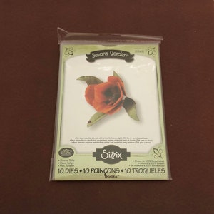 May include: A Sizzix Thinlits die set for crafting, featuring a die-cut red tulip with green leaves. The package displays the text "Susan's Garden" and "Sizzix". The set contains 10 dies for creating floral designs.