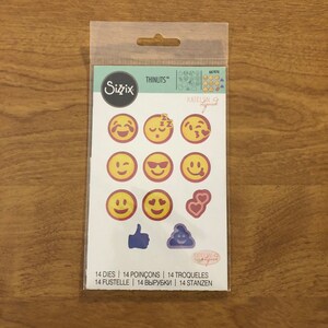 May include: A Sizzix Thinlits die set with emoji designs, including smiling faces, hearts, a thumbs up, and a poo emoji. The package is labelled with the text "14 DIES" and is in a clear plastic wrapper.