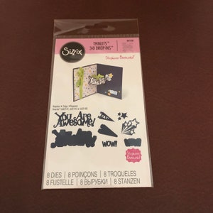 May include: A packet of Sizzix Thinlits 3-D Drop-Ins die cuts. The package contains various die cuts for making greeting cards, including the words "You Are Awesome!" and starburst shapes. The package is sealed in clear plastic.
