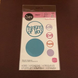 May include: A Sizzix Thinlits die set featuring sentiments like "thinking of you", "hello", "love", and "thanks", plus two circles. The package, designed by Stephanie Burnard, includes six dies. The die-cuts are in a variety of colours.