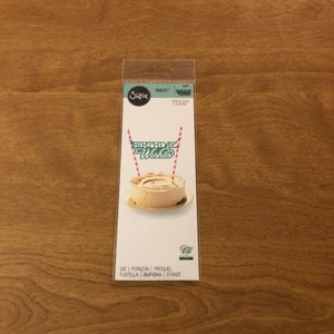 May include: A Sizzix Thinlits die cut package for cake decoration. The card displays "Birthday Wishes" in green script, with two pink and white striped straws. A frosted pastry is shown on a plate. The package is on a wooden surface.