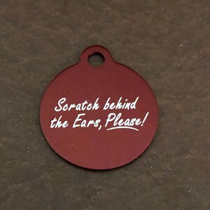 Peut inclure: Une médaille pour animal de compagnie circulaire bordeaux avec un trou en haut. Le texte blanc sur la médaille dit : "Scratch behind the Ears, Please !" La médaille est probablement en métal et est conçue pour être attachée au collier d'un animal.