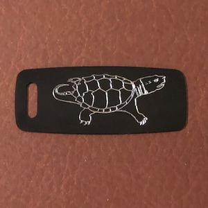 May include: Black luggage tag featuring a silver turtle design. The tag is rectangular with rounded corners and a slot for attachment. The turtle is shown with a detailed shell, walking to the right.