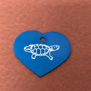 May include: A blue heart-shaped tag featuring a white etched turtle design. The tag has a small hole at the top for hanging. The turtle is drawn in a simple, illustrative style.