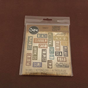 May include: A pack of Sizzix Thinlits dies featuring travel-themed words such as "Beach," "Summer," and "Vacation." The die-cut shapes are in various colours and sizes, ideal for scrapbooking or card making.