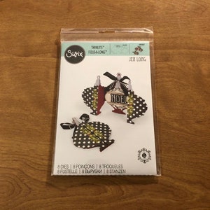 May include: A pack of Sizzix Thinlits Fold-A-Long dies for making Christmas decorations. The dies are designed to create two different ornaments, one with a "NOEL" inscription and the other with a polka dot pattern. The pack contains 8 dies.
