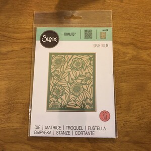 May include: A Sizzix Thinlits die set with a floral design. The die is a rectangle with a detailed pattern of flowers and leaves. The packaging includes the text "SOPHIE GUILAR" and "DIE | MATRICE | TROQUEL | FUSTELLA".