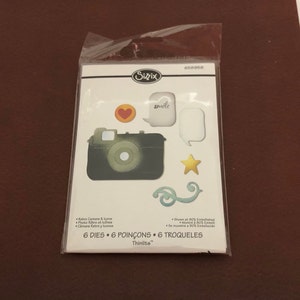 May include: A Sizzix Thinlits die set with retro camera and icon designs. The set includes a camera, heart, star, speech bubbles, and decorative swirls. The package is sealed in clear plastic.