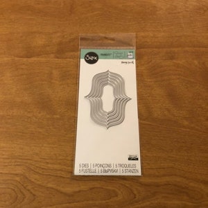 May include: A packet of Sizzix Framelits dies, featuring a nested bracket design. The dies are silver and come in a clear plastic package with text in multiple languages. The package includes 5 dies.