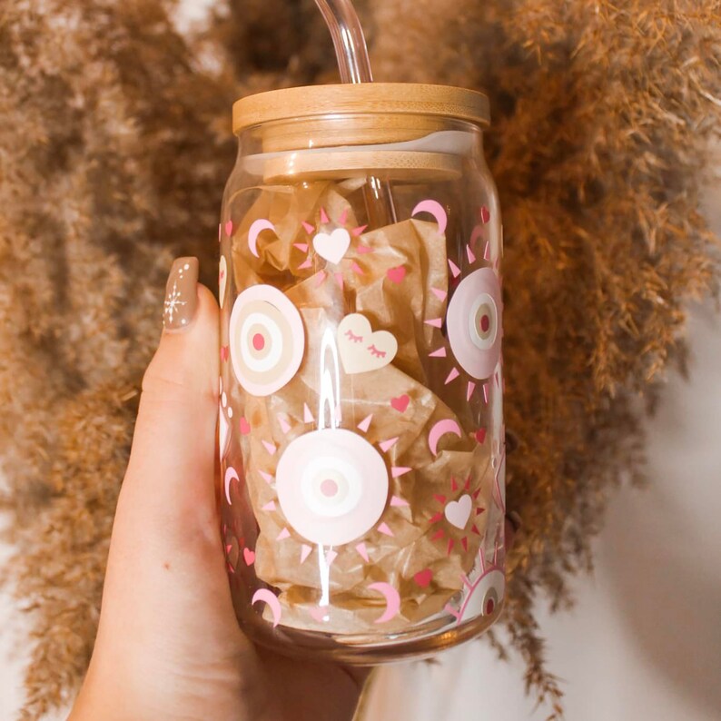 May include: A glass reusable water bottle with a bamboo lid and a straw. The bottle has a pink and white design with eyes, hearts, and suns.