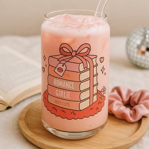 May include: A pink glass can with a straw, filled with a pink beverage. The can has a graphic of a stack of books tied with a pink ribbon, with the words "Born", "Romance", "Girlie", and "Meet Cute" on the books.