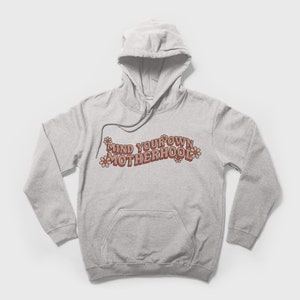 May include: A gray hooded sweatshirt with the text "MIND YOUR OWN MOTHERHOOD" in pink and brown letters with pink flowers on either side of the text.