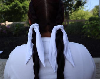 May include: A rear view of a person with long, dark braided hair. Each braid is tied with a white fabric. The person is wearing a white shirt, with a blurred green background.