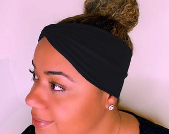 May include: A black headband is worn across the forehead. The headband is made of a soft, stretchy material. The hair is styled in a bun. The background is a solid light pink color. The person is wearing a black top and a silver necklace.