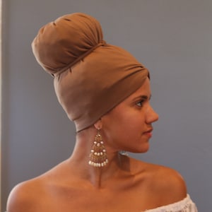 Puede incluir: Una mujer con un turbante marrón de estilo turbante con un gran moño en la parte superior. Lleva grandes pendientes colgantes dorados y blancos.