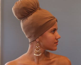 Puede incluir: Una mujer con un turbante marrón de estilo turbante con un gran moño en la parte superior. Lleva grandes pendientes colgantes dorados y blancos.