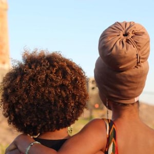 May include: Two people, one with brown curly hair and the other with a brown turban, are standing close together. The person with the turban is wearing a brown sleeveless dress.