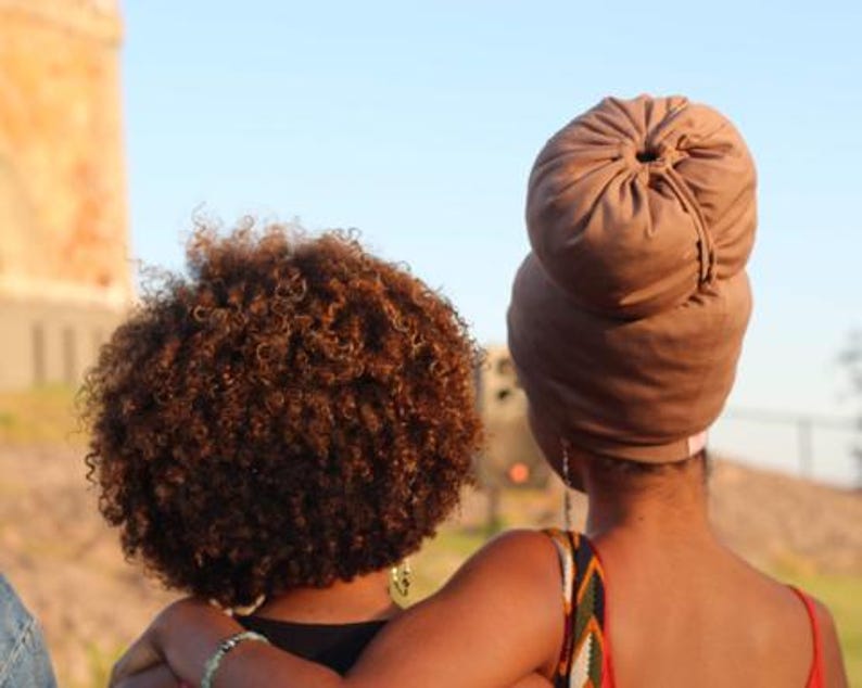 May include: Two people, one with brown curly hair and the other with a brown turban, are standing close together. The person with the turban is wearing a brown sleeveless dress.