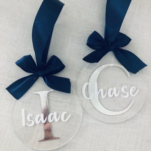 May include: Two clear acrylic ornaments with navy blue ribbon bows. One ornament has the name "Isaac" with a copper-colored "I", and the other says "Chase" with a white "C". Both have navy blue ribbon loops.