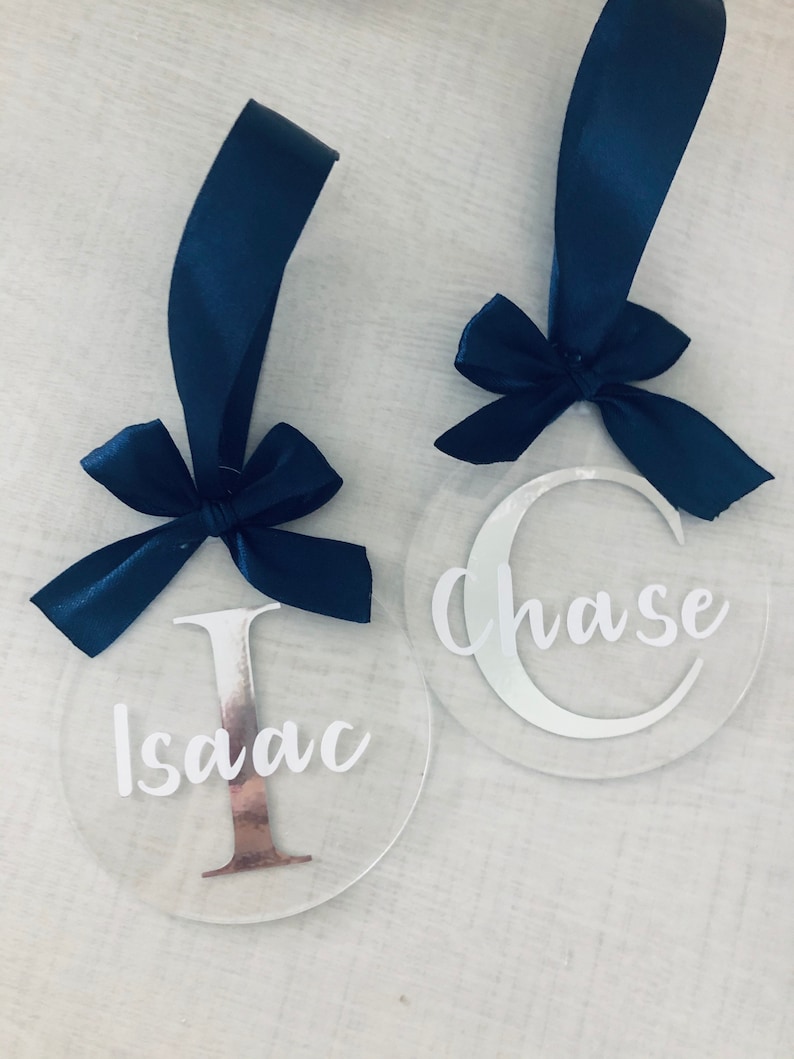 May include: Two clear acrylic ornaments with navy blue ribbon bows. One ornament has the name "Isaac" with a copper-colored "I", and the other says "Chase" with a white "C". Both have navy blue ribbon loops.