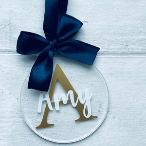 May include: A clear, circular ornament with the name "Amy" in white script and a gold letter "A". A navy blue ribbon is attached at the top. The ornament is hanging against a white wood-grain background.