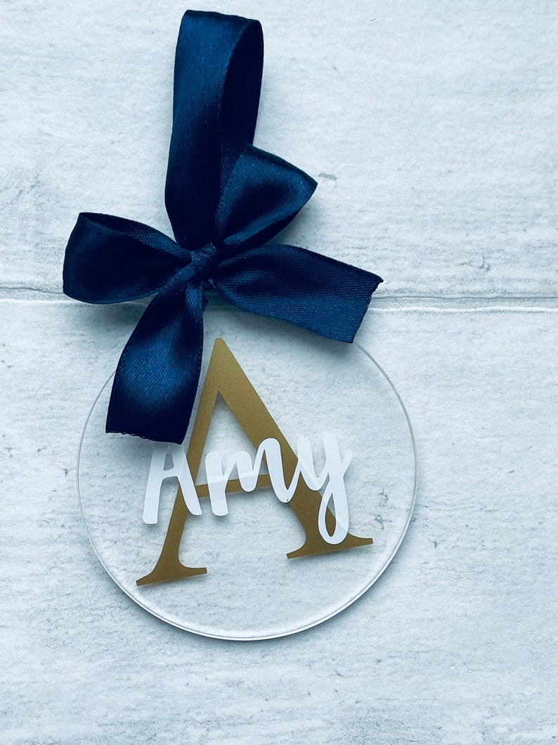 May include: A clear, circular ornament with the name "Amy" in white script and a gold letter "A". A navy blue ribbon is attached at the top. The ornament is hanging against a white wood-grain background.
