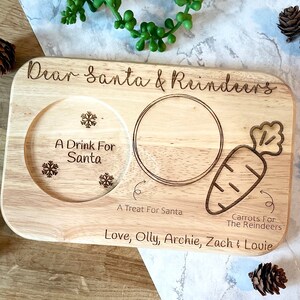 May include: Wooden serving tray with a carved design for Santa and reindeer. It has a place for a drink, a treat, and carrots. The tray is personalized with the text "Dear Santa & Reindeers", "A Drink For Santa", "A Treat For Santa", "Carrots For The Reindeers", and "Love, Olly, Archie, Zach & Lovie".