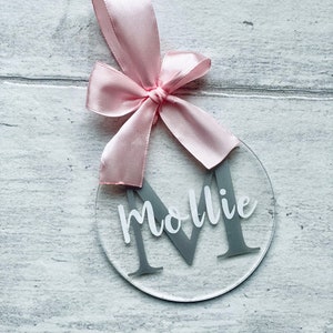 May include: Clear acrylic ornament with a gray letter "M" and the name "Mollie" in white script. A pink satin ribbon is tied at the top in a bow for hanging. The ornament is round and sits on a light gray wooden surface.