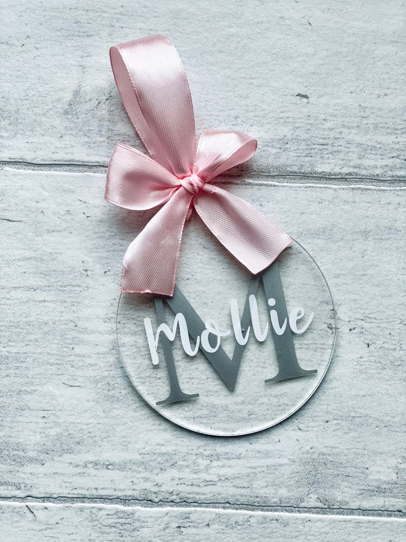 May include: Clear acrylic ornament with a gray letter "M" and the name "Mollie" in white script. A pink satin ribbon is tied at the top in a bow for hanging. The ornament is round and sits on a light gray wooden surface.