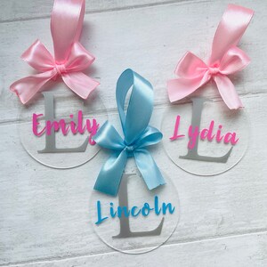 May include: Three clear acrylic circle ornaments with personalized names and initial letters. Each ornament has a satin ribbon bow in pink or blue. The names "Emily" and "Lydia" are in pink, and "Lincoln" is in blue.