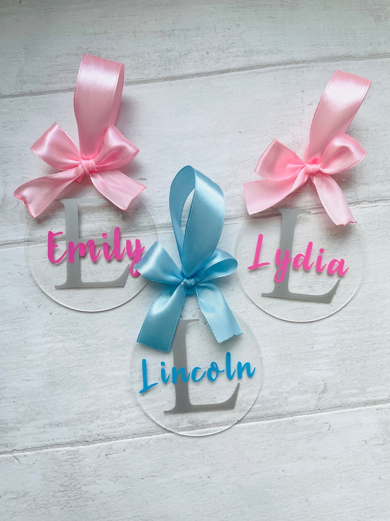 May include: Three clear acrylic circle ornaments with personalized names and initial letters. Each ornament has a satin ribbon bow in pink or blue. The names "Emily" and "Lydia" are in pink, and "Lincoln" is in blue.