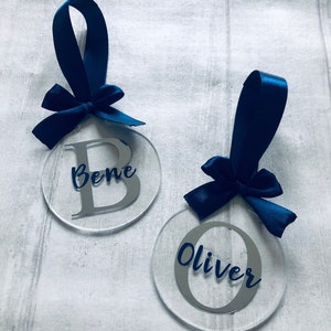 May include: Two clear acrylic ornaments with navy blue satin ribbon bows. One ornament has the name "Bene" with a silver letter "B", and the other has the name "Oliver" with a silver letter "O".