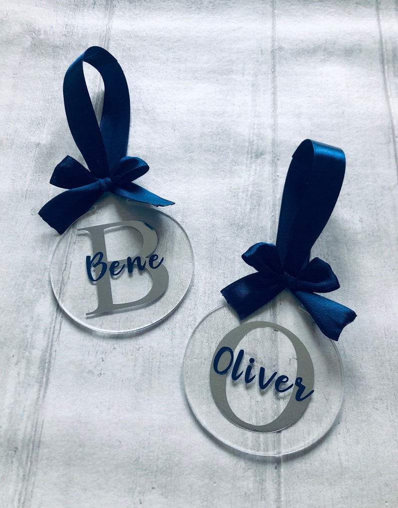 May include: Two clear acrylic ornaments with navy blue satin ribbon bows. One ornament has the name "Bene" with a silver letter "B", and the other has the name "Oliver" with a silver letter "O".