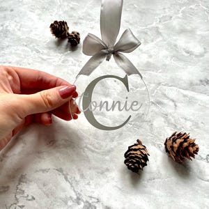 May include: Clear acrylic Christmas ornament with the name "Connie" in gray script. The ornament has a gray ribbon and bow for hanging. Three small pinecones are scattered around the ornament on a marble surface.