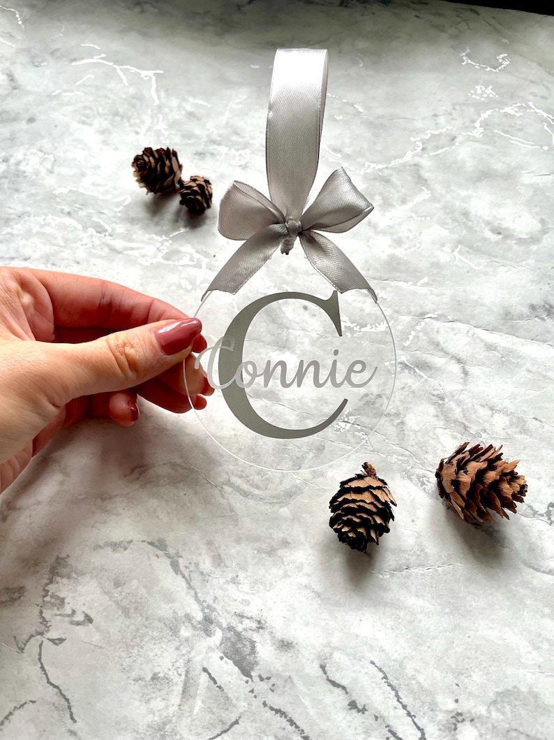 May include: Clear acrylic Christmas ornament with the name "Connie" in gray script. The ornament has a gray ribbon and bow for hanging. Three small pinecones are scattered around the ornament on a marble surface.