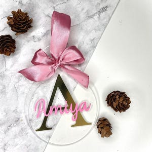 May include: A clear, round ornament with the name "Amaya" in pink script, set over a gold letter "A". A pink ribbon is attached at the top. The ornament is surrounded by pinecones.