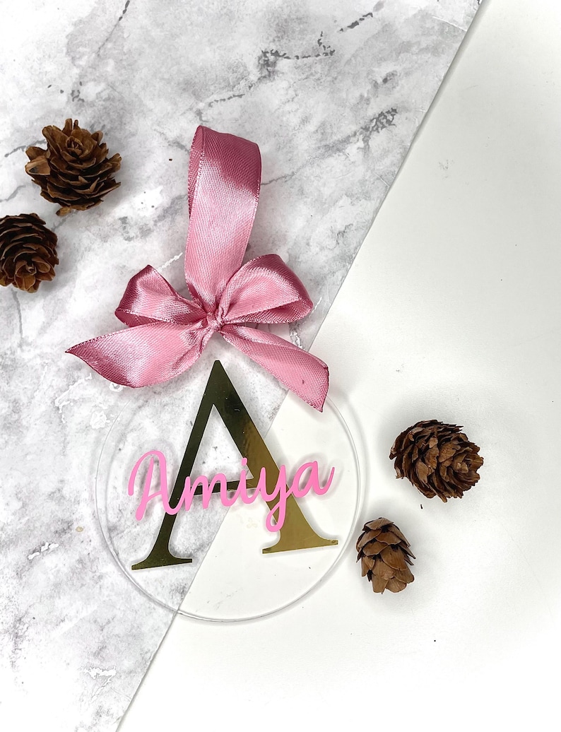 May include: A clear, round ornament with the name "Amaya" in pink script, set over a gold letter "A". A pink ribbon is attached at the top. The ornament is surrounded by pinecones.
