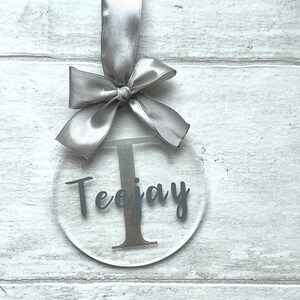 May include: Clear acrylic ornament with the name "Teejay" and the letter "T" in a dark gray font. The ornament is suspended by a silver satin ribbon tied in a bow.