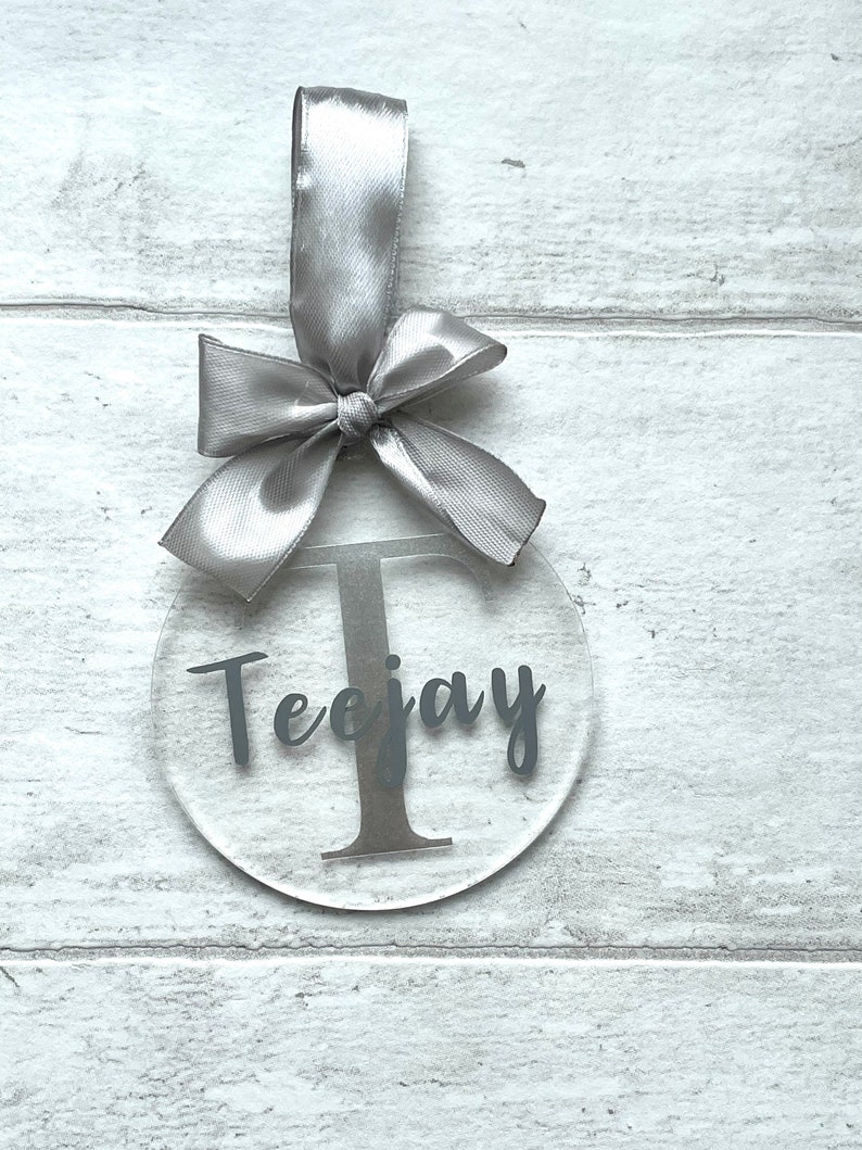 May include: Clear acrylic ornament with the name "Teejay" and the letter "T" in a dark gray font. The ornament is suspended by a silver satin ribbon tied in a bow.