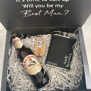 May include: A gift box with the text "It's time to suit up, Will you be my Best Man?" contains a beer bottle, a flask, and a bottle opener. The flask is black with the name "Andy Bestman" and the date "28.05.2025".