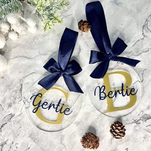 May include: Two clear, round Christmas ornaments with navy blue ribbon bows. One ornament has a gold "G" and the name "Gertie" in navy script. The other has a gold "B" and the name "Bertie" in navy script. Pinecones and greenery are in the background.