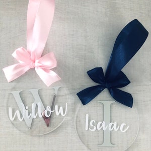 May include: Two clear, round ornaments with names, "Willow" and "Isaac", in white script. Each ornament has a satin ribbon and bow. One ribbon is pink, the other is navy blue. The ornaments are likely for a special occasion.