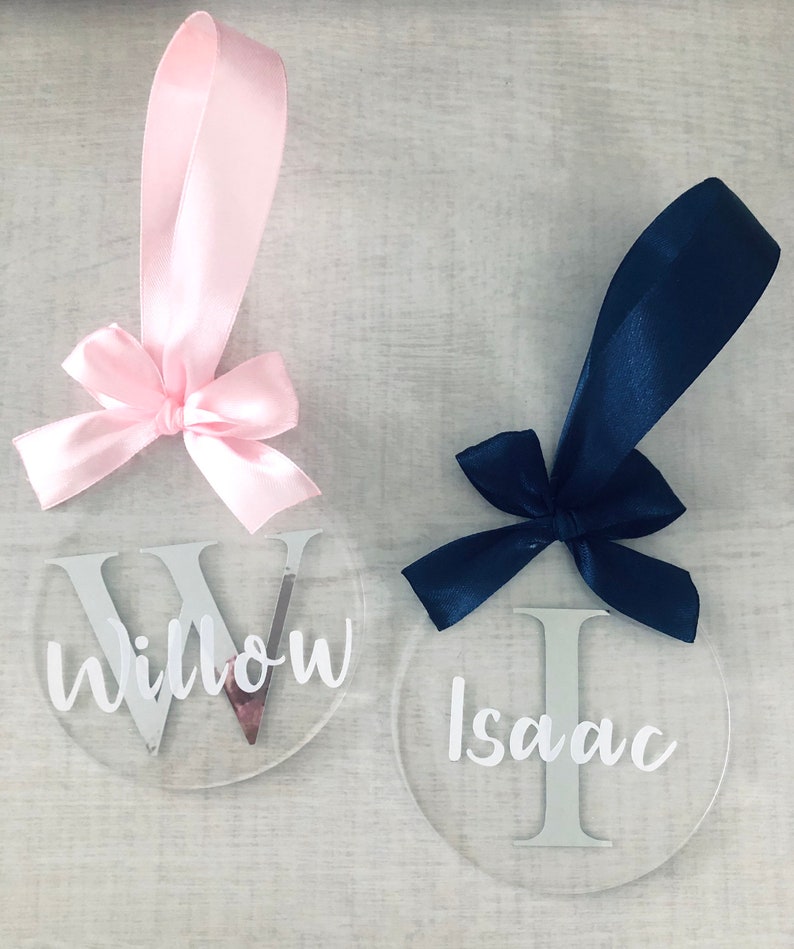 May include: Two clear, round ornaments with names, "Willow" and "Isaac", in white script. Each ornament has a satin ribbon and bow. One ribbon is pink, the other is navy blue. The ornaments are likely for a special occasion.